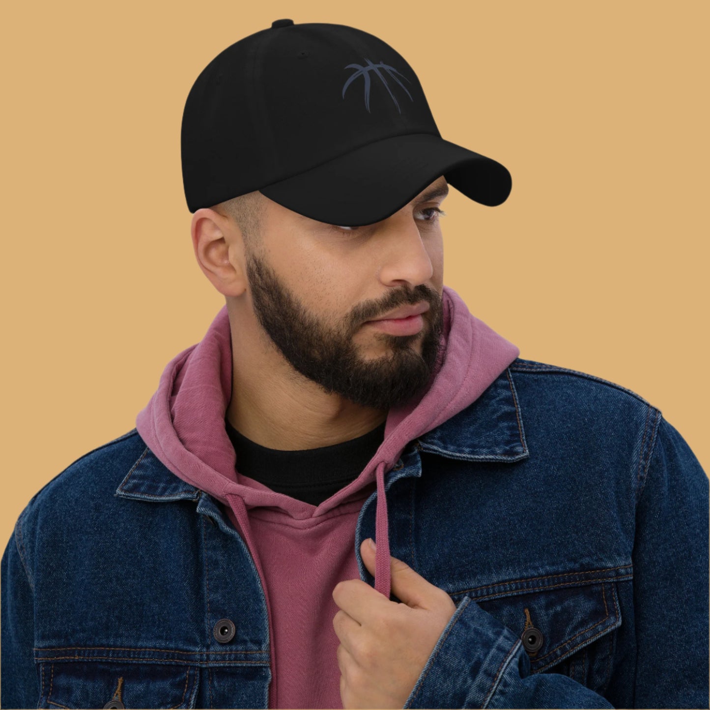 Man wearing a black cap with a logo, denim jacket, and pink hoodie against a gray background