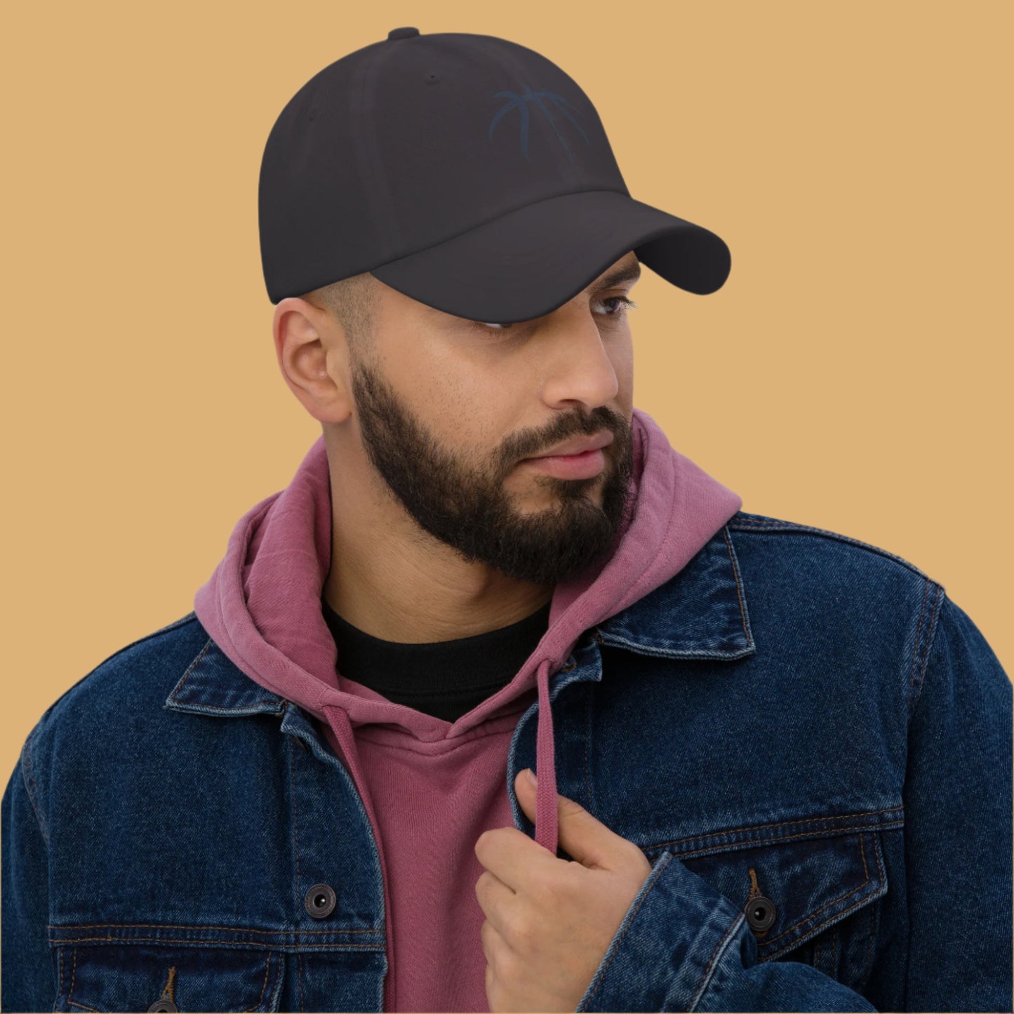Man wearing a black cap, pink hoodie, and blue denim jacket against a gray background