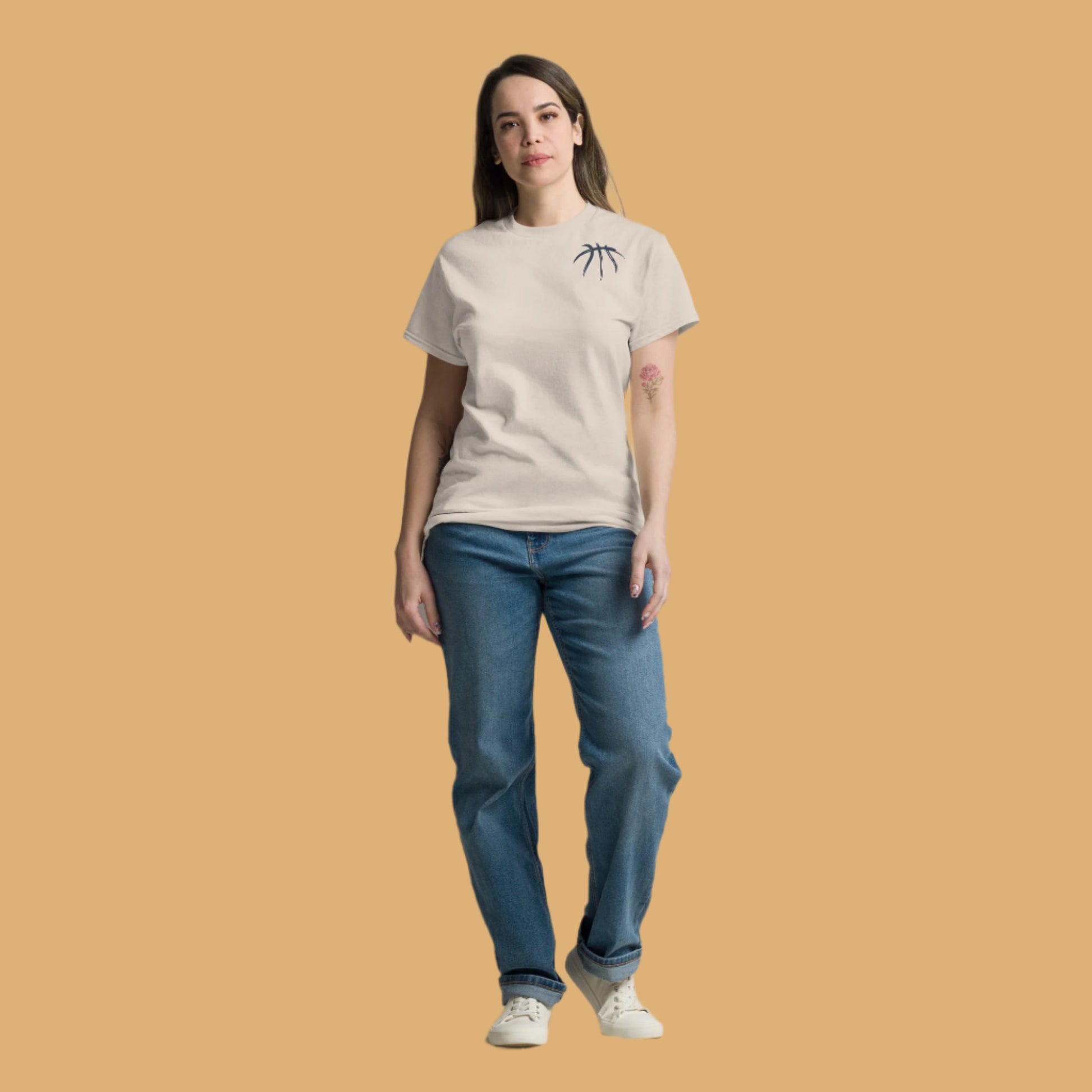 Woman wearing a beige t-shirt and blue jeans standing against a plain white curtain.