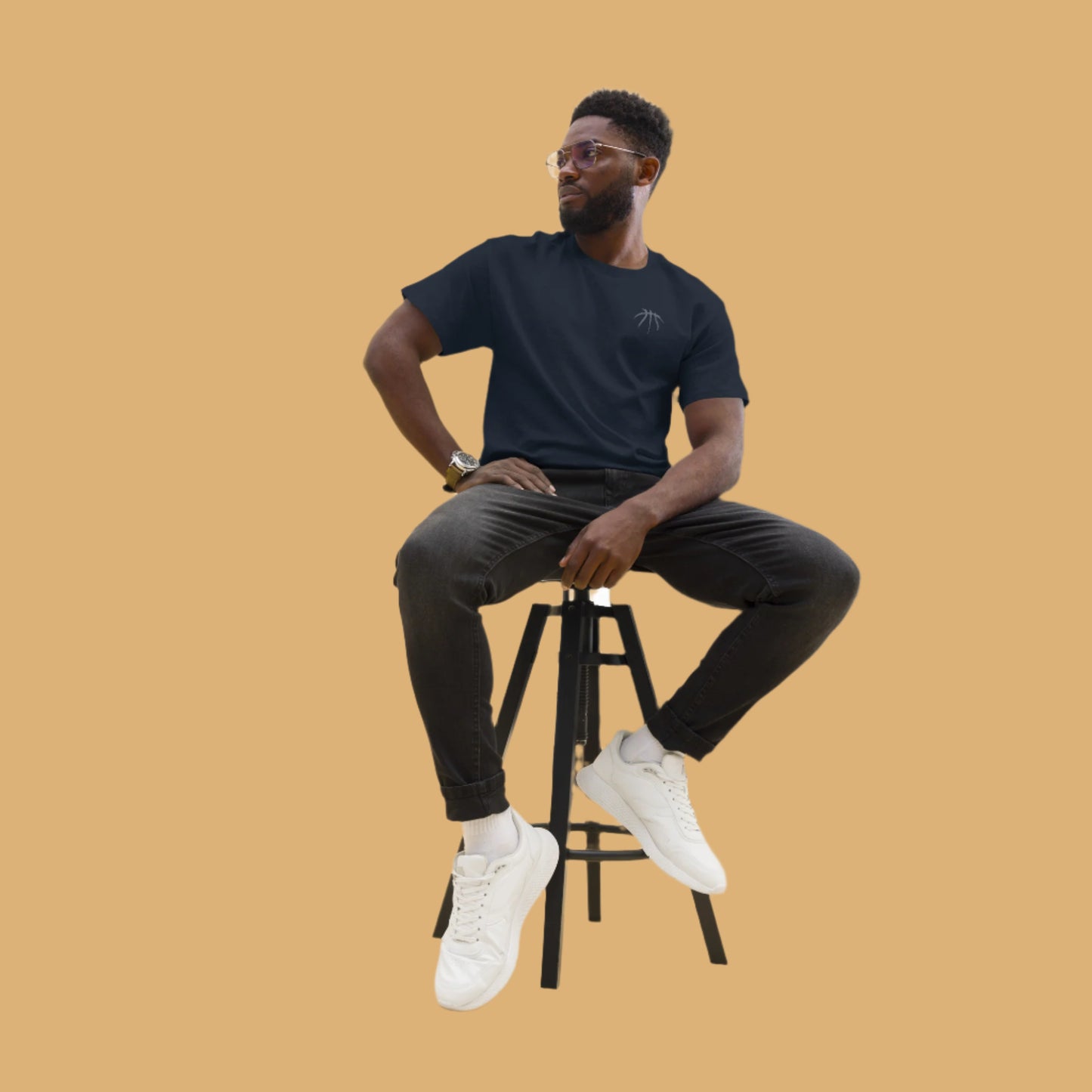Man sitting on a stool wearing a black t-shirt, black pants, and white sneakers against a beige background