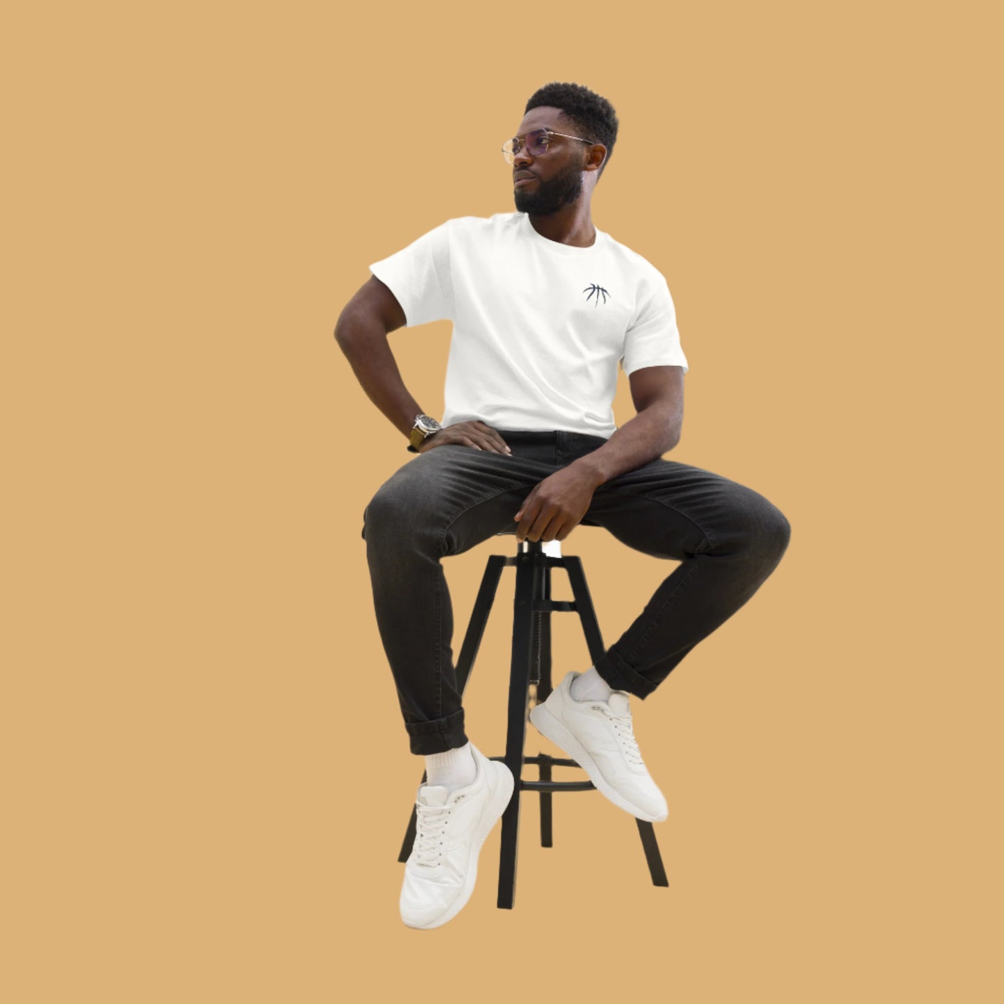 Man sitting on a stool wearing a white t-shirt, black pants, and white sneakers against a plain background
